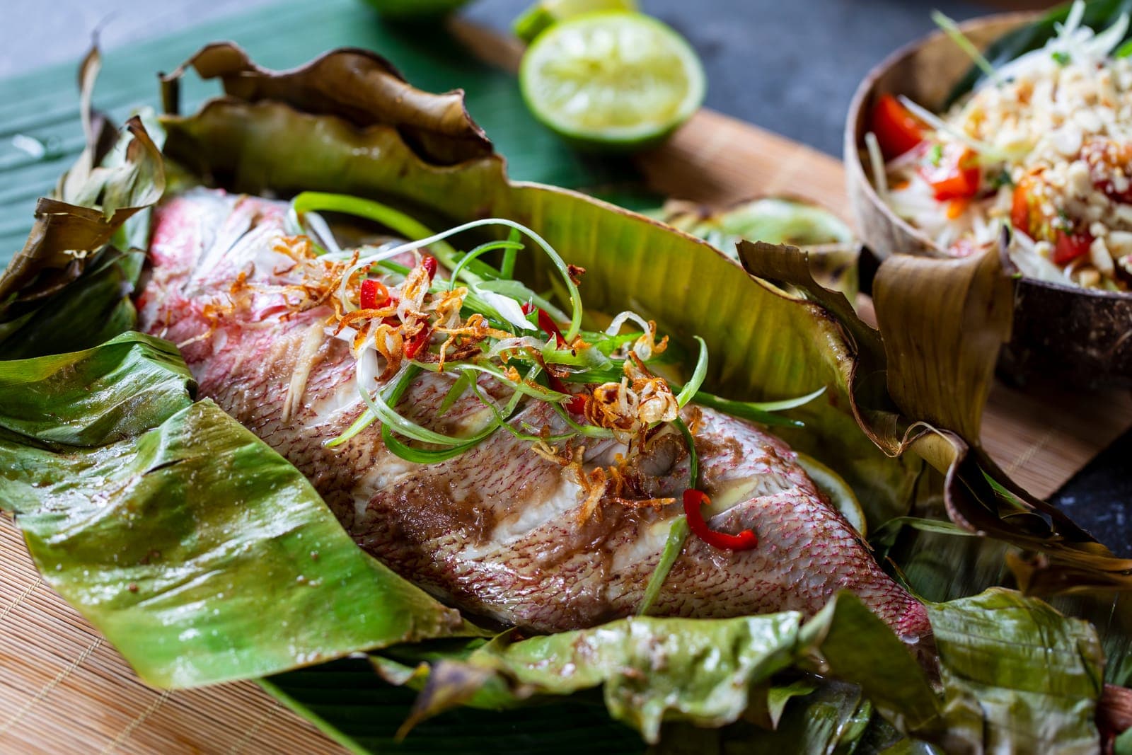 A whole fish grilled in a banana leaf parcel, opened to show the flesh with ginger, scallion, and lime